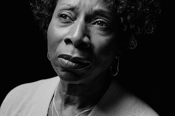 Portrait en noir et blanc d'une femme âgée à l'air pensif, le regard légèrement levé, sur un fond sombre, son visage éclairé par la droite.